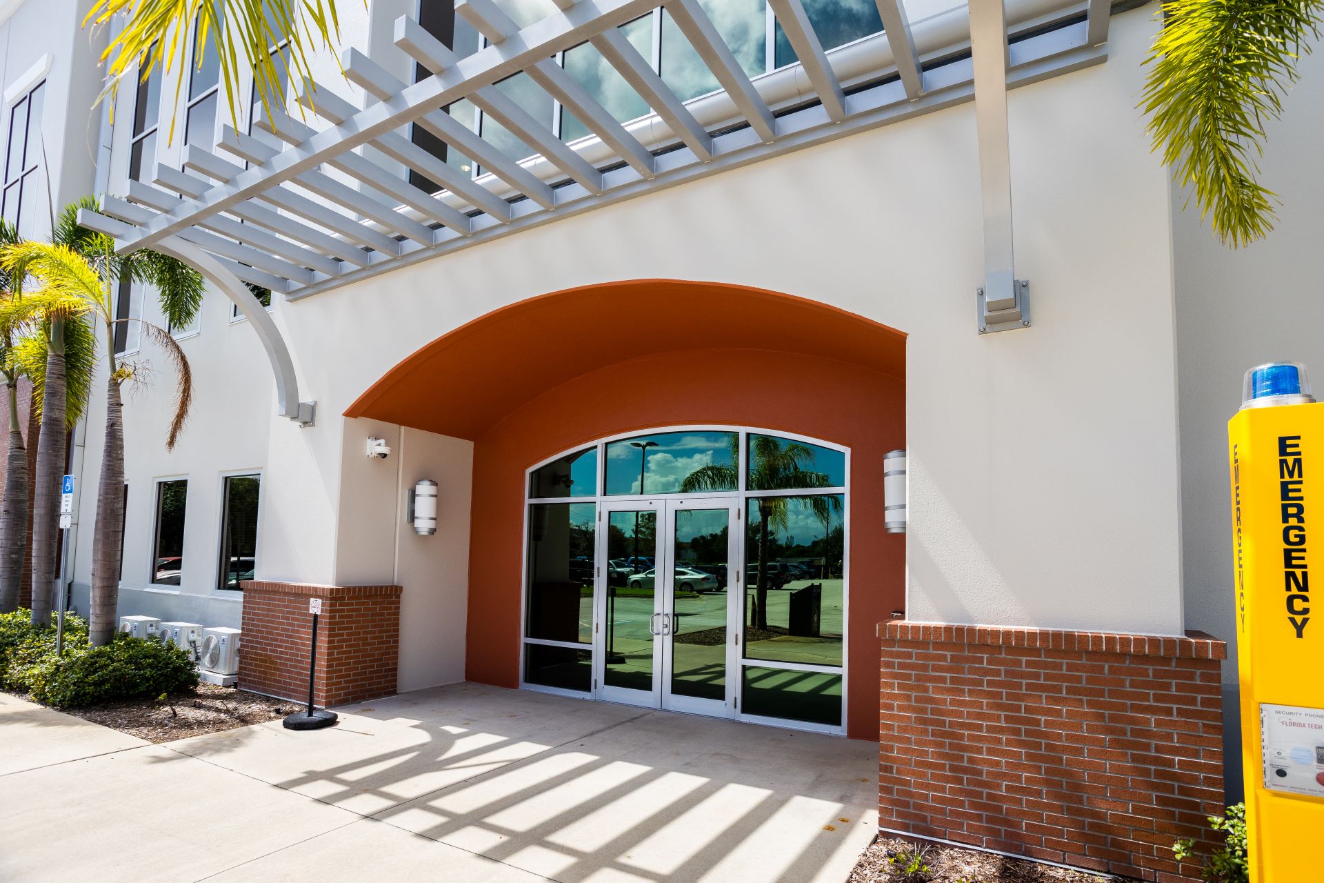 Close-up view of the main entrance of the L3Harris Commons at Florida Tech, highlighting the modern design with large glass doors, an arched entryway, and an emergency station on the right, set against a backdrop of palm trees and clear skies